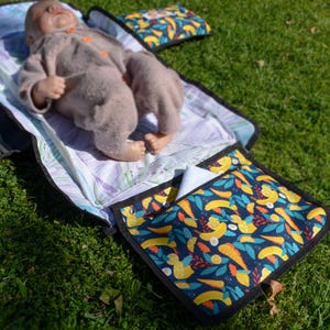 May include: A baby changing station with a colorful pattern of yellow birds, orange carrots, and green leaves on a navy blue background. The changing pad is open, with a baby doll lying on it. The changing station is on a green grassy surface.