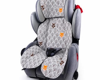 May include: A gray car seat featuring a repeating pattern of stylized trees interspersed with orange and brown fox faces. The seat includes adjustable side headrests, a central harness buckle, and textured gray fabric on the outer side panels.