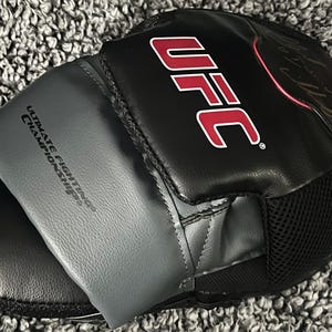 Puede incluir: Guante de entrenamiento de artes marciales mixtas UFC negro y gris. El guante presenta el logotipo de la UFC en rojo y blanco, con las palabras "Ultimate Fighting Championships" impresas en la correa de la muñeca. El guante tiene un panel de malla para ventilación.