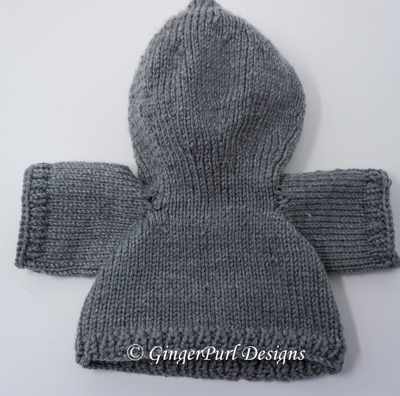 Puede incluir: Un su&eacute;ter con capucha gris tejido con mangas. El su&eacute;ter est&aacute; hecho de un hilo grueso y tiene un dobladillo y pu&ntilde;os acanalados. La capucha tiene punta en la parte superior. El texto "&copy; GingerPurl Designs" es visible en la parte inferior.