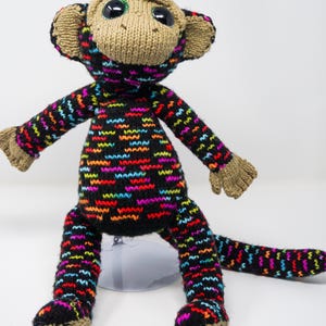 Puede incluir: Mu&ntilde;eco de mono tejido a mano con cuerpo y extremidades negros, adornado con un colorido patr&oacute;n en zigzag en rojo, azul, amarillo y morado. El mono tiene cara, orejas, manos y pies color beige, y ojos grandes y expresivos.