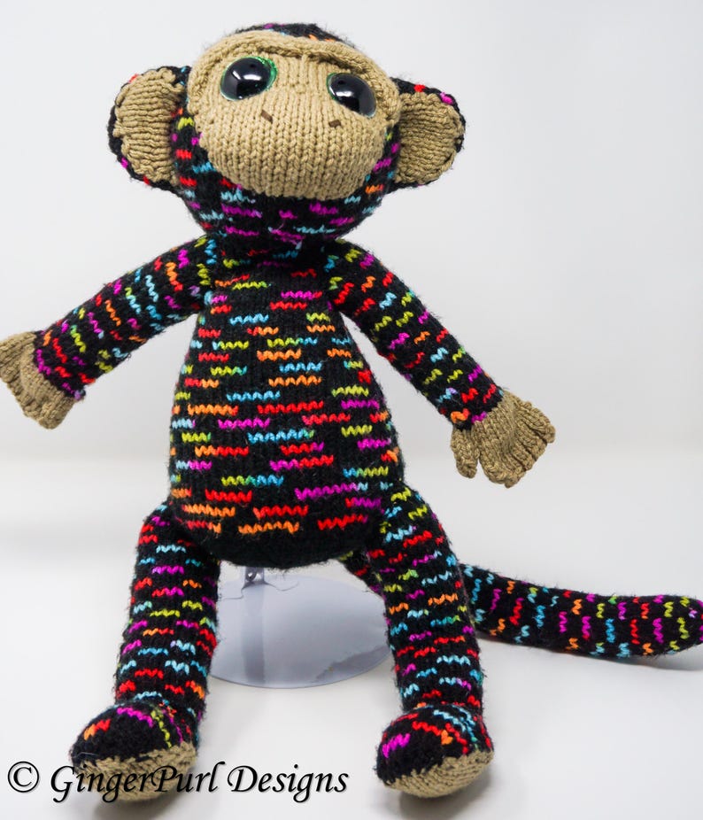 Puede incluir: Mu&ntilde;eco de mono tejido a mano con cuerpo y extremidades negros, adornado con un colorido patr&oacute;n en zigzag en rojo, azul, amarillo y morado. El mono tiene cara, orejas, manos y pies color beige, y ojos grandes y expresivos.