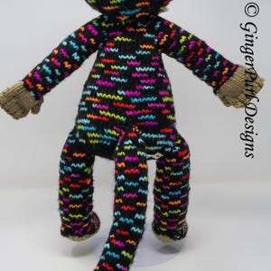 Puede incluir: Mu&ntilde;eco de mono tejido a mano con un fondo negro y patrones de zigzag coloridos en rojo, naranja, amarillo, verde, azul y morado. El mono tiene manos y pies marrones. La mu&ntilde;eca est&aacute; sobre una superficie blanca.