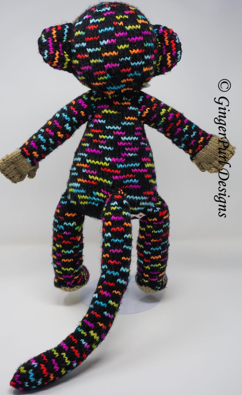 Puede incluir: Mu&ntilde;eco de mono tejido a mano con un fondo negro y patrones de zigzag coloridos en rojo, naranja, amarillo, verde, azul y morado. El mono tiene manos y pies marrones. La mu&ntilde;eca est&aacute; sobre una superficie blanca.