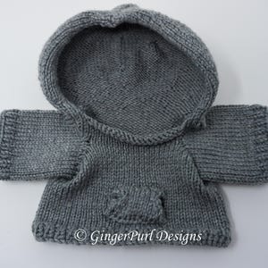 Puede incluir: Una sudadera con capucha gris tejida con capucha y bolsillo delantero. El su&eacute;ter hecho a mano tiene mangas largas y est&aacute; dise&ntilde;ado para un animal peque&ntilde;o o una mu&ntilde;eca. El texto "GingerPurl Designs" es visible en la parte inferior.
