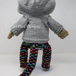 Puede incluir: Mu&ntilde;eca tejida a mano con un su&eacute;ter con capucha gris, pantalones estampados y guantes color canela. La mu&ntilde;eca tiene una cola larga y est&aacute; sobre una base blanca. Los pantalones son negros con patrones de zigzag coloridos.