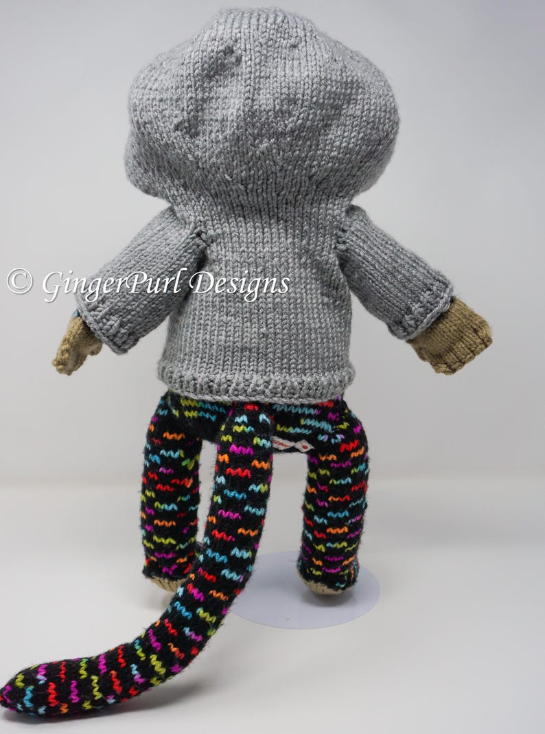 Puede incluir: Mu&ntilde;eca tejida a mano con un su&eacute;ter con capucha gris, pantalones estampados y guantes color canela. La mu&ntilde;eca tiene una cola larga y est&aacute; sobre una base blanca. Los pantalones son negros con patrones de zigzag coloridos.
