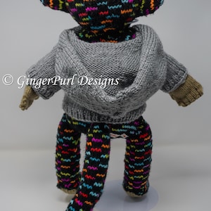 Puede incluir: Un mu&ntilde;eco de mono tejido a mano con una sudadera con capucha gris y pantalones estampados. La cabeza y los pantalones del mono tienen un fondo negro con patrones de zigzag coloridos. El mu&ntilde;eco tiene manos y pies de color beige.