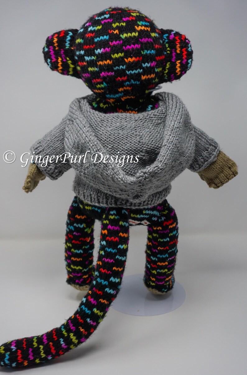 Puede incluir: Un mu&ntilde;eco de mono tejido a mano con una sudadera con capucha gris y pantalones estampados. La cabeza y los pantalones del mono tienen un fondo negro con patrones de zigzag coloridos. El mu&ntilde;eco tiene manos y pies de color beige.