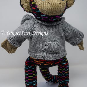 Puede incluir: Muñeco de mono tejido a mano con grandes ojos brillantes. El mono lleva un suéter gris con bolsillo y pantalones negros con un patrón ondulado y colorido. La cabeza y las orejas del mono son de color canela, con un gorro negro y colorido.