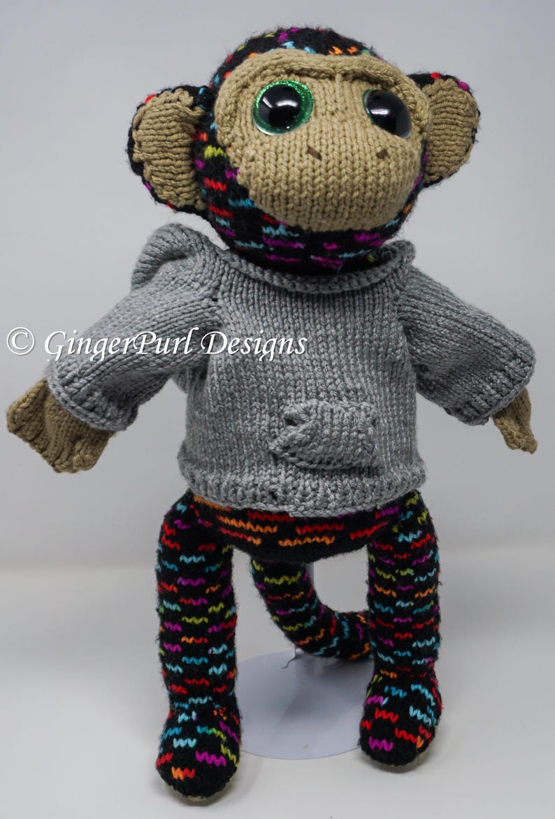 Puede incluir: Mu&ntilde;eco de mono tejido a mano con grandes ojos brillantes. El mono lleva un su&eacute;ter gris con bolsillo y pantalones negros con un patr&oacute;n ondulado y colorido. La cabeza y las orejas del mono son de color canela, con un gorro negro y colorido.