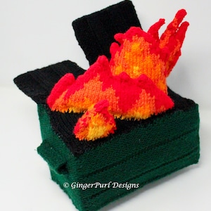 May include: A knitted sculpture of a green skip with a black lid and flames. The flames are red, orange, and yellow, and appear to be coming out of the skip. The skip is made of a knitted material.