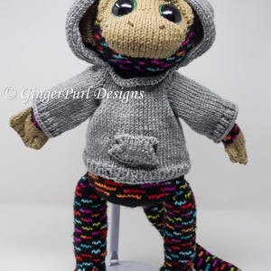 Puede incluir: Mu&ntilde;eca de mono tejida a mano con un su&eacute;ter con capucha gris, una bufanda colorida y leggings estampados. La mu&ntilde;eca tiene ojos negros grandes y est&aacute; sobre un soporte blanco. El texto "GingerPurl Designs" es visible.