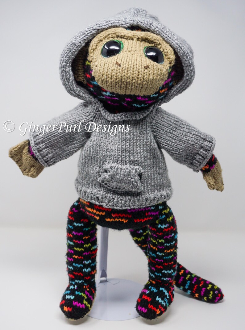 Puede incluir: Mu&ntilde;eca de mono tejida a mano con un su&eacute;ter con capucha gris, una bufanda colorida y leggings estampados. La mu&ntilde;eca tiene ojos negros grandes y est&aacute; sobre un soporte blanco. El texto "GingerPurl Designs" es visible.