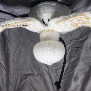May include: A handmade white knitted angel doll with gold detailing on the wings. The doll has a round body and head, with a black string and a green tassel. The wings are spread, and the text "GingerPurl Designs" is visible.