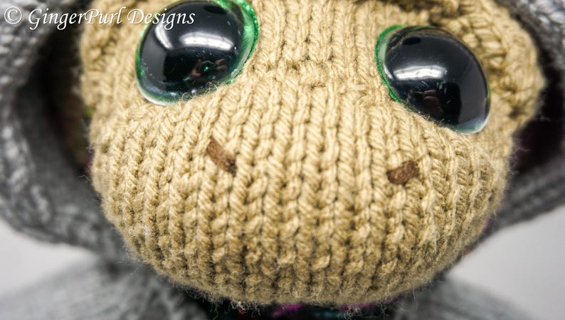 Puede incluir: Primer plano de una cabeza de mu&ntilde;eca tejida con ojos negros grandes y brillantes y una cara de color canela. La mu&ntilde;eca tiene un su&eacute;ter con capucha gris. El texto "GingerPurl Designs" est&aacute; en la esquina superior izquierda.