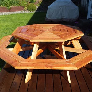 K&ouml;nnte beinhalten: Achteckiger Holz-Picknicktisch mit B&auml;nken aus nat&uuml;rlichem Zedernholz. Der Tisch ist in einem satten Braunton gebeizt und hat eine glatte Oberfl&auml;che. Der Tisch steht auf einem Holzdeck.