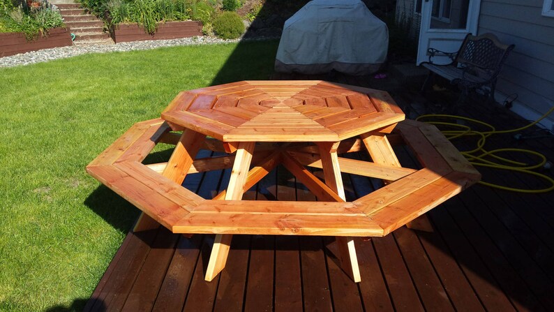 K&ouml;nnte beinhalten: Achteckiger Holz-Picknicktisch mit B&auml;nken aus nat&uuml;rlichem Zedernholz. Der Tisch ist in einem satten Braunton gebeizt und hat eine glatte Oberfl&auml;che. Der Tisch steht auf einem Holzdeck.