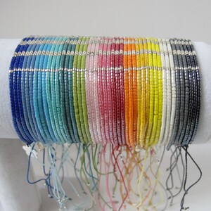 Può includere: Una collezione di braccialetti colorati con perline e dettagli argentati, con chiusura regolabile in corda. I braccialetti sono disposti a forma di arcobaleno, con ogni braccialetto che presenta un colore diverso.