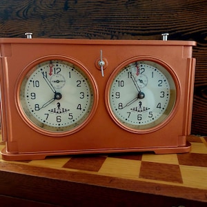 May include: A vintage orange chess clock with two analog faces. Each face has black numbers and hands, and a small white dial with black markings. The clock is on a wooden chessboard.