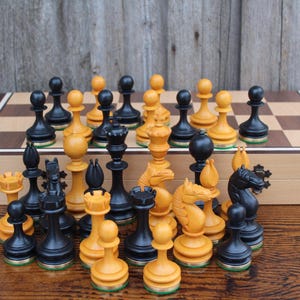 Può includere: Un set di scacchi completo con pezzi neri e in legno chiaro disposti su una scacchiera di legno. I pezzi includono pedoni, torri, cavalli, alfieri, una regina e un re. La scacchiera è posizionata su una superficie di legno.