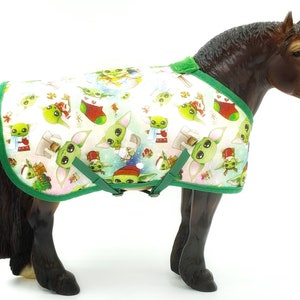 Peut inclure: Un mod&egrave;le de cheval portant une couverture blanche avec une bordure verte. La couverture est orn&eacute;e de personnages de dessins anim&eacute;s verts et blancs, de bas de No&euml;l rouges et de flocons de neige. La couverture a des sangles et des boucles vertes.