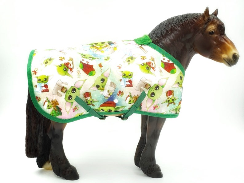 Peut inclure: Un mod&egrave;le de cheval portant une couverture blanche avec une bordure verte. La couverture est orn&eacute;e de personnages de dessins anim&eacute;s verts et blancs, de bas de No&euml;l rouges et de flocons de neige. La couverture a des sangles et des boucles vertes.