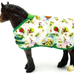 Peut inclure: Une couverture pour cheval avec un motif r&eacute;p&eacute;t&eacute; d'un personnage extraterrestre vert et d'&eacute;l&eacute;ments de vacances festifs. La couverture a un fond blanc avec une bordure et des sangles vertes. Le cheval est de couleur marron fonc&eacute;.
