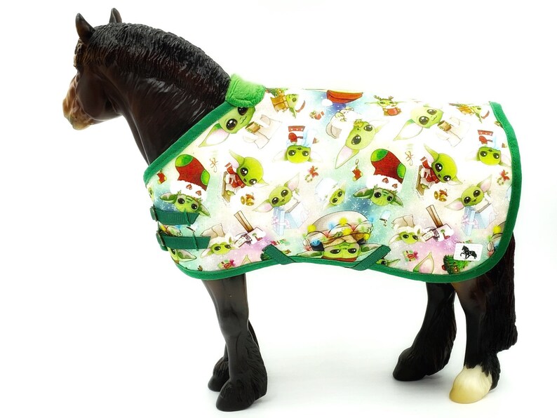 Peut inclure: Une couverture pour cheval avec un motif r&eacute;p&eacute;t&eacute; d'un personnage extraterrestre vert et d'&eacute;l&eacute;ments de vacances festifs. La couverture a un fond blanc avec une bordure et des sangles vertes. Le cheval est de couleur marron fonc&eacute;.