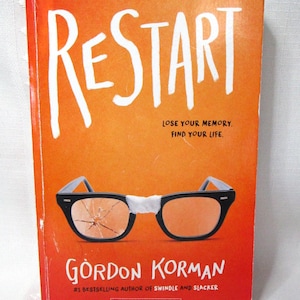 May include: A book cover with the title "RESTART" in white letters on an orange background. Below the title are the words "LOSE YOUR MEMORY. FIND YOUR LIFE." and an illustration of broken glasses. The author's name, Gordon Korman, is also displayed.
