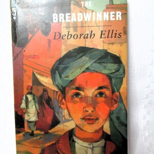 May include: A book cover for "The Breadwinner" by Deborah Ellis. The cover features a portrait of a young person wearing a teal turban and a red jacket. The background shows a street scene with figures.