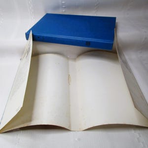 Puede incluir: Un libro azul de tapa dura descansa sobre un libro abierto con p&aacute;ginas envejecidas de color blanquecino. El libro abierto est&aacute; colocado para crear una forma de tienda de campa&ntilde;a, con el libro azul como &aacute;pice. Las p&aacute;ginas muestran signos de edad.