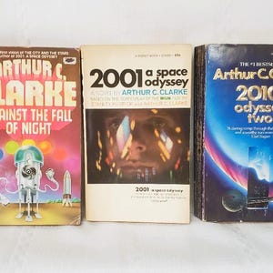 May include: Three vintage science fiction books by Arthur C. Clarke. Titles include "Against the Fall of Night," "2001: A Space Odyssey," and "2010: Odyssey Two." The books have colorful covers with retro designs and text.