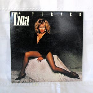 May include: Album cover for Tina Turner's "Private Dancer." The album cover features a glamorous portrait of Tina Turner in a black outfit, with the word "Tina" in white block letters at the top. The background is dark.