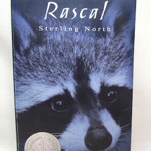 May include: A book cover featuring a close-up of a raccoon's face with the title "Rascal" and author "Sterling North" in white text. A silver coin is at the bottom. The background is dark blue.