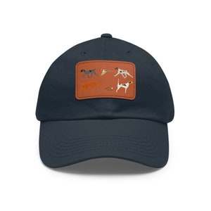 Op de afbeelding: Een donkerblauwe baseballpet met een bruine leren patch met illustraties van jachthonden en vogels.