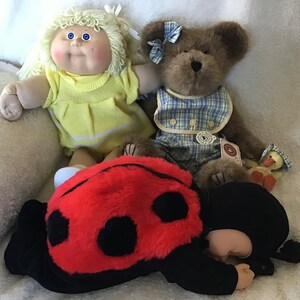 May include: A plush ladybug toy with black spots and red fur, a blonde doll with blue eyes and a yellow dress, and a brown teddy bear wearing a blue and white plaid dress.