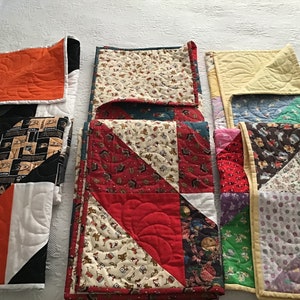 May include: A collection of nine folded quilts in various colors and patterns, including orange, black, white, red, green, blue, yellow, and purple. The quilts are made of different fabrics and have a variety of quilting designs.