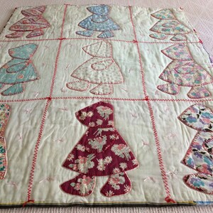 May include: A vintage quilt with a patchwork design featuring nine appliquéd figures of girls wearing bonnets. The figures are made from various floral fabrics in shades of pink, blue, red, and purple. The quilt is stitched with red and pink thread.