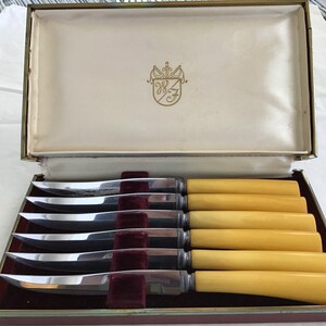 May include: Set of six silver-plated steak knives with yellow plastic handles in a burgundy velvet-lined box. The box has a gold trim and a crest with the initials "W&F" in gold.