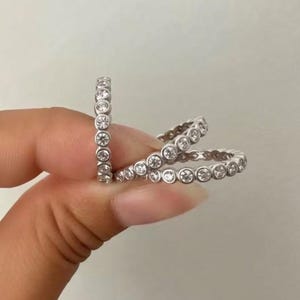 May include: A set of three silver-toned eternity rings, each featuring a continuous band of small, round, clear stones set in a bezel style. The rings are arranged in a cluster, showcasing the metallic finish and the circular stone settings.