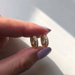 May include: A pair of gold hoop earrings with a row of clear cubic zirconia stones around the edge.