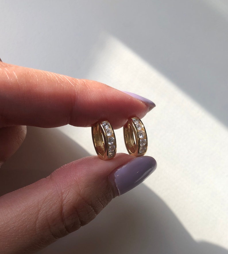 May include: A pair of gold hoop earrings with a row of clear cubic zirconia stones around the edge.