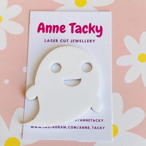 May include: A white ghost-shaped laser-cut jewelry piece with a smiling face, displayed on a white card with the text "Anne Tacky LASER CUT JEWELLERY". The background is pink with white daisy patterns.