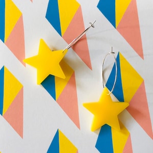 May include: A pair of yellow star-shaped earrings with silver hoops. The earrings are on a white background with a geometric pattern of pink, blue, and yellow triangles.