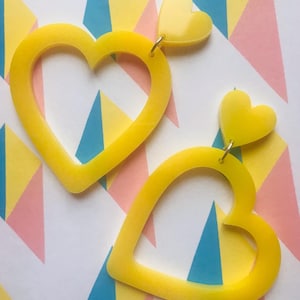 May include: A pair of yellow heart-shaped earrings with a smaller heart charm hanging from each. The earrings are made of a smooth, glossy material.
