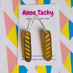 May include: Gold-coloured earrings shaped like a baguette with a swirl pattern. The earrings are on a white card with the text "Anne Tacky Laser Cut Jewellery" and website information.