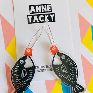 May include: Fish-shaped earrings with a black design and silver hoops. The earrings are suspended in front of a white card with the text "ANNE TACKY" in a black box. The background features a colourful geometric pattern.
