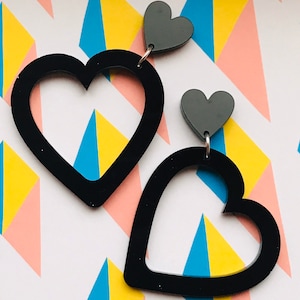 May include: A pair of black heart-shaped earrings with a smaller black heart charm hanging from each. The earrings are on a white background with a geometric pattern.