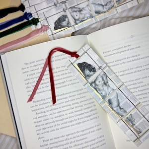 May include: A handmade bookmark featuring a grayscale illustration of a nude figure, set against a backdrop of book pages. The bookmark has a red ribbon and is surrounded by other velvet ribbon bookmarks.